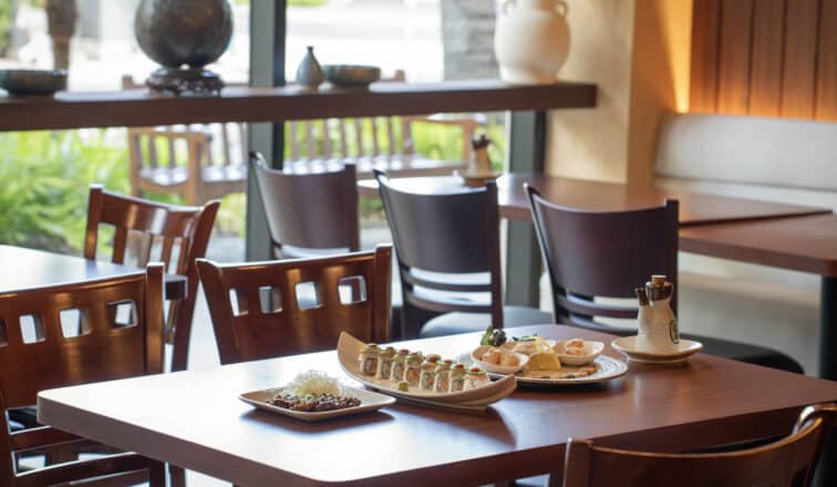 A restaurant table set with plates of sushi and appetizers, with empty wooden chairs and large windows in the background.