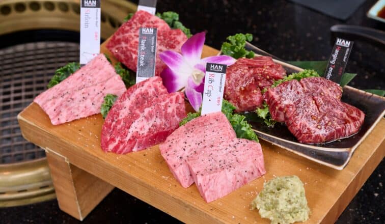 A wooden tray displays various cuts of raw marbled beef labeled with tags, garnished with greens, an orchid flower, and a dollop of wasabi, next to a grill.