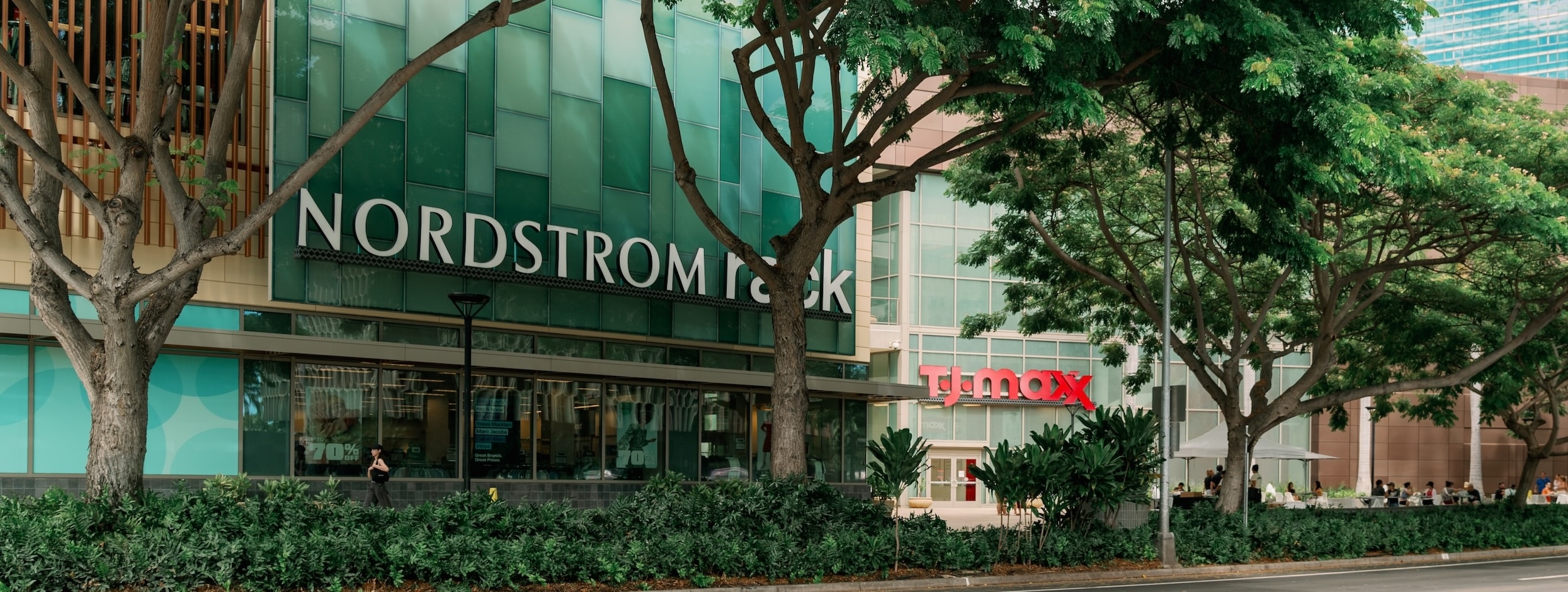 Nordstrom Rack and TJ Maxx storefronts are visible behind trees and greenery in an urban shopping area.