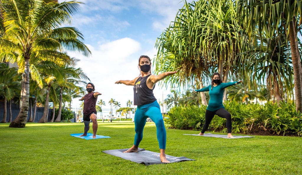 Two athletic women and one man doing yoga in the park wearing face masks