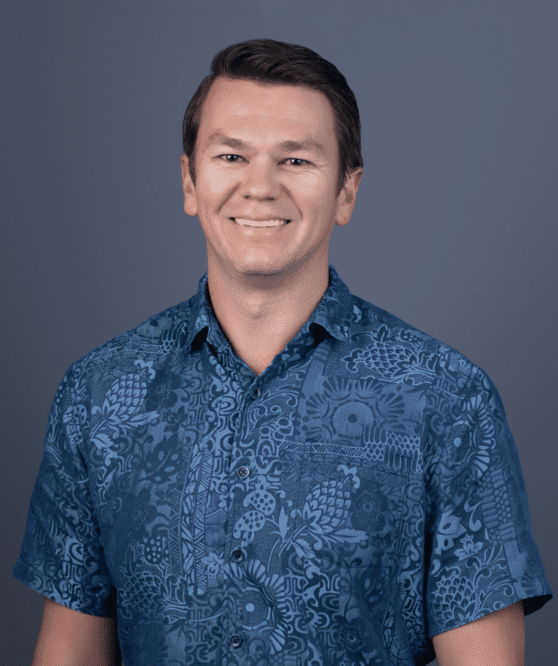 A man with short brown hair wearing a blue patterned short-sleeve shirt stands against a plain blue background, smiling at the camera.