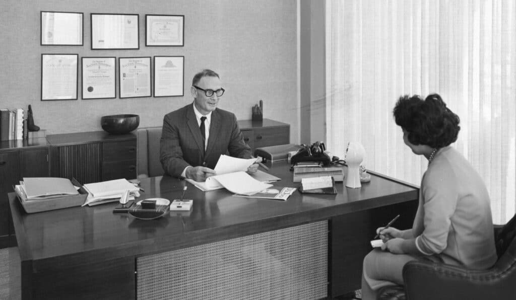 Inside the IBM Building, a man sits behind a desk and a woman in a chair is facing him with a note pad.