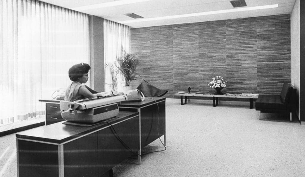 Inside the IBM Building a woman sits behind a desk with a typewriter and mid-century office decor.
