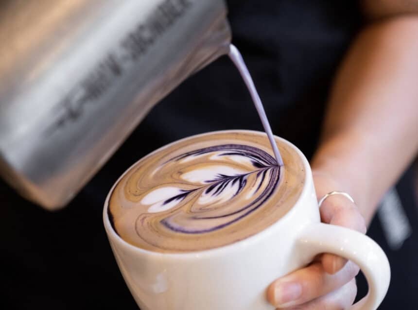 Close-up action shot of a barista pouring cream into a cup of coffee creating a purple flower design