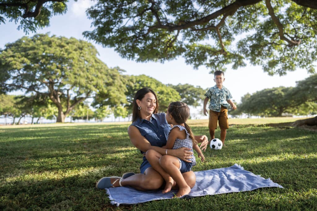 A family at the park with a woman holding a child while sitting on a blanket on the ground