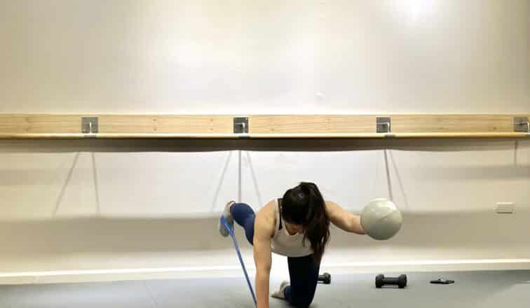 A person exercises indoors on a mat, balancing on one knee with a resistance band and holding a gray ball, with a dumbbell and other equipment nearby.
