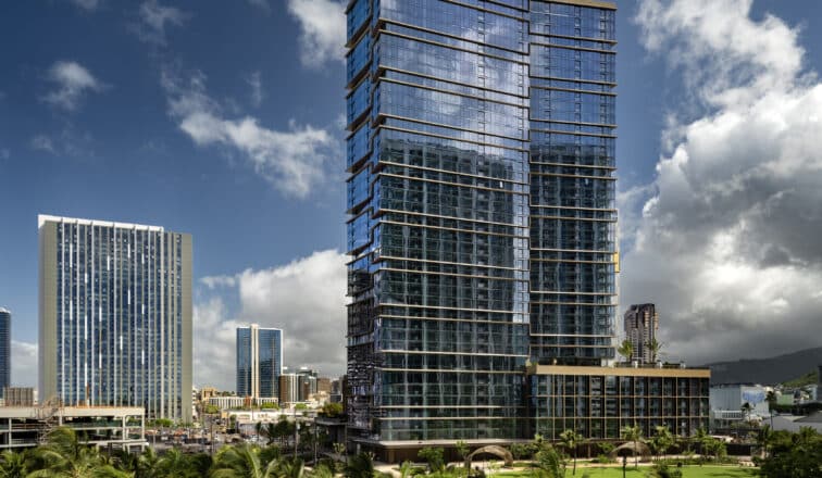 A tall, modern glass skyscraper stands amid other high-rise buildings and palm trees under a partly cloudy sky.