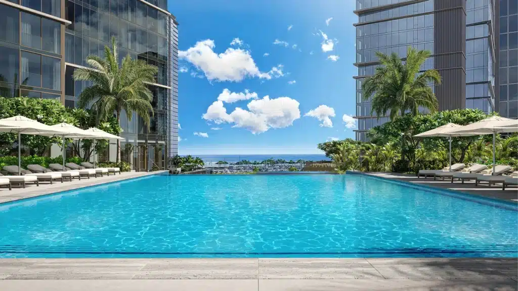 Large outdoor swimming pool surrounded by lounge chairs and palm trees, situated between modern high-rise buildings, with a view of the ocean in the background under a blue sky.