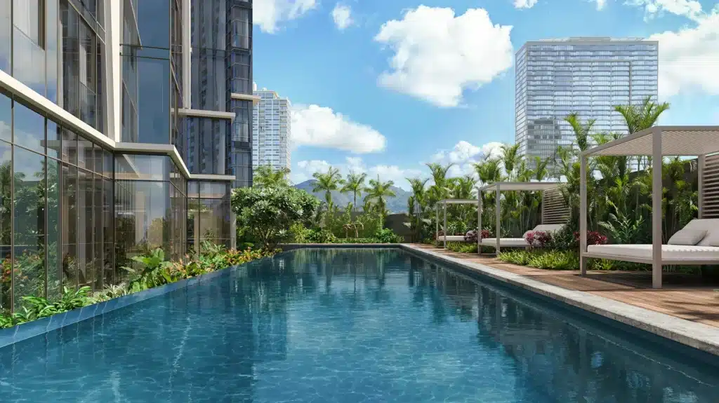 Rooftop swimming pool with clear water, surrounded by plants and lounge cabanas, adjacent to tall modern buildings under a blue sky with clouds.