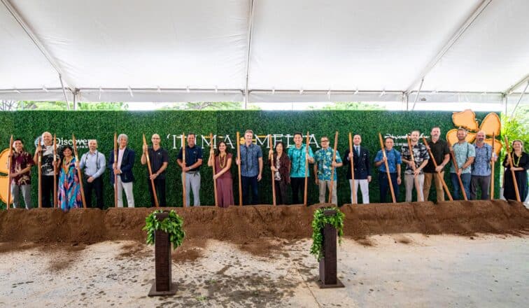 A group of people stand in a line holding shovels at a groundbreaking ceremony under a large white tent, with a green hedge and event signage behind them.