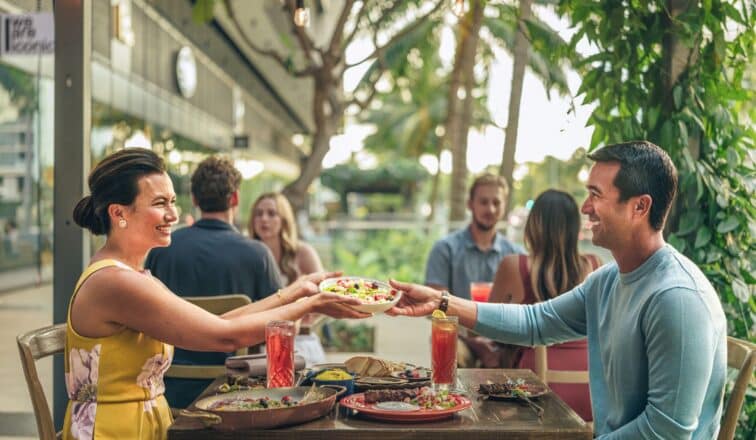 A woman and man sit at an outdoor restaurant table, smiling and sharing a plate of food, with other diners and lush greenery in the background.