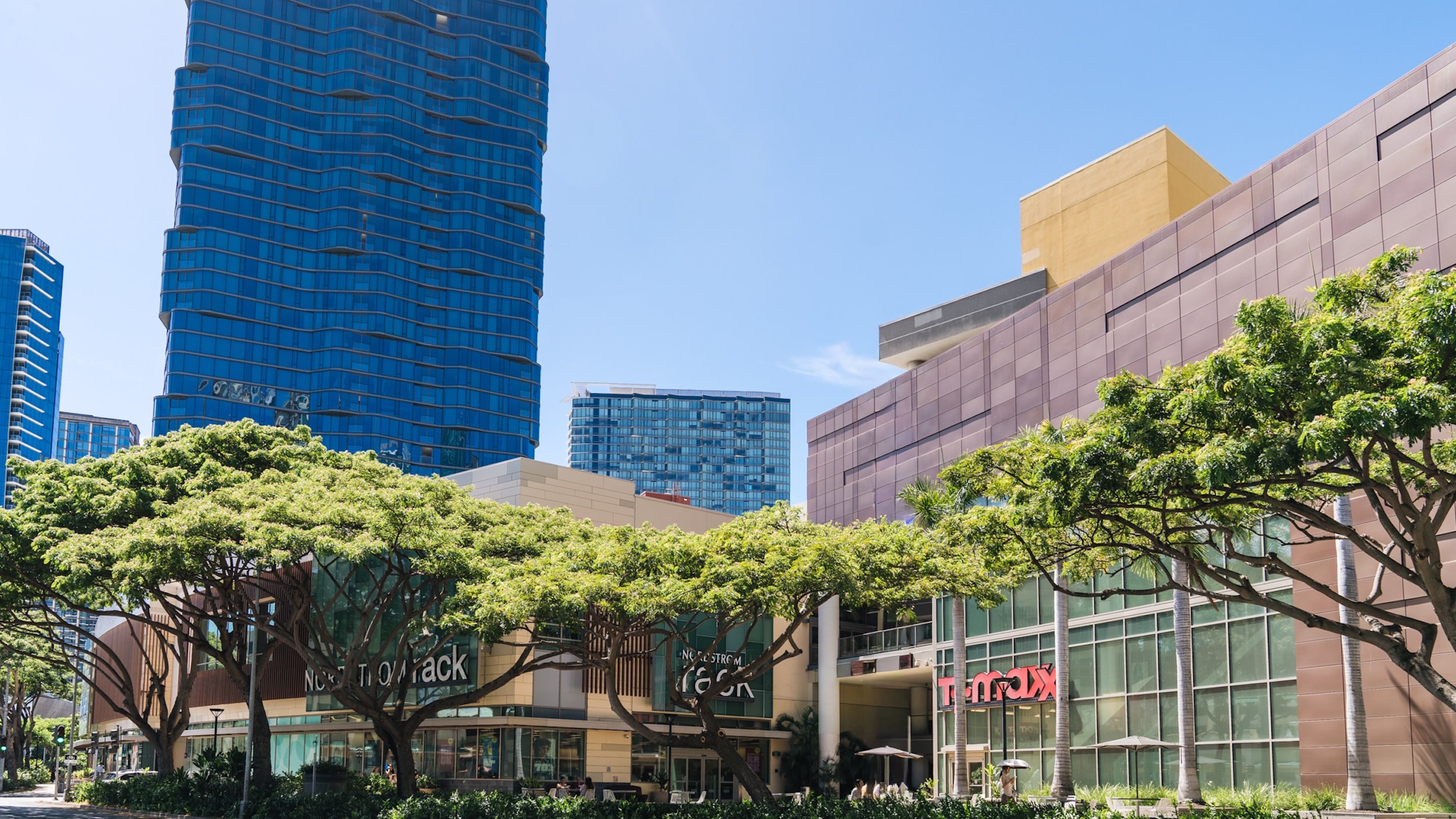 Nordstrom Rack and TJ Maxx storefronts with tall modern buildings and leafy green trees under a clear blue sky.